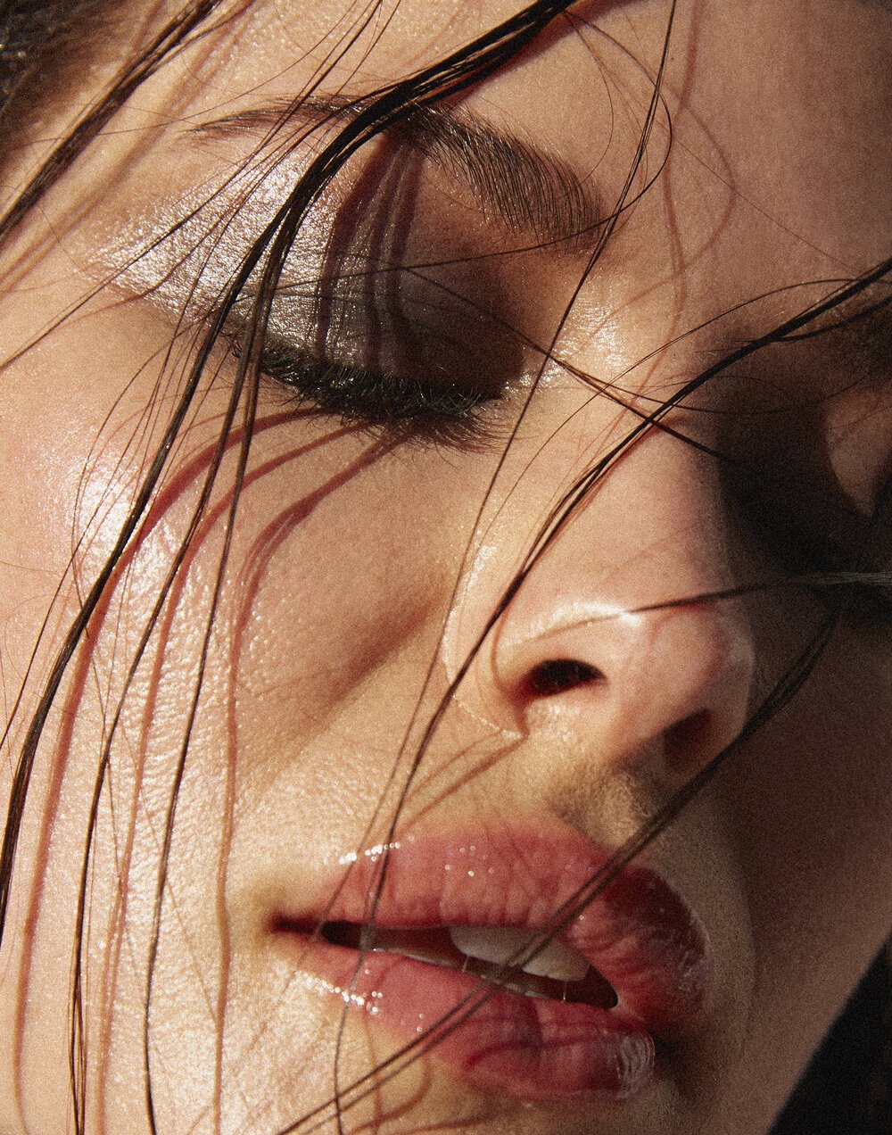 Close-up portrait of a woman with her eyes closed, glossy lips, and wet dark hair framing her face in soft sunlight.