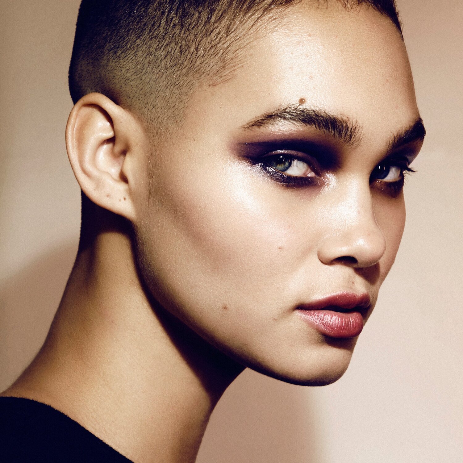 Close-up portrait of a person with short, dark hair, vibrant purple eyeshadow, and a neutral background.