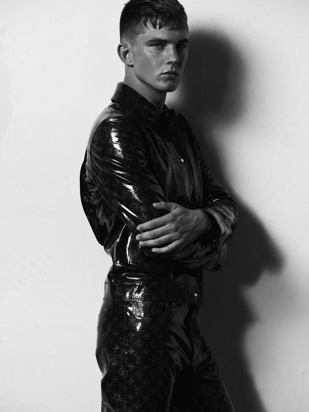 Monochrome fashion portrait of a young man in a shiny black jacket and patterned pants, posing against a plain white wall backdrop.