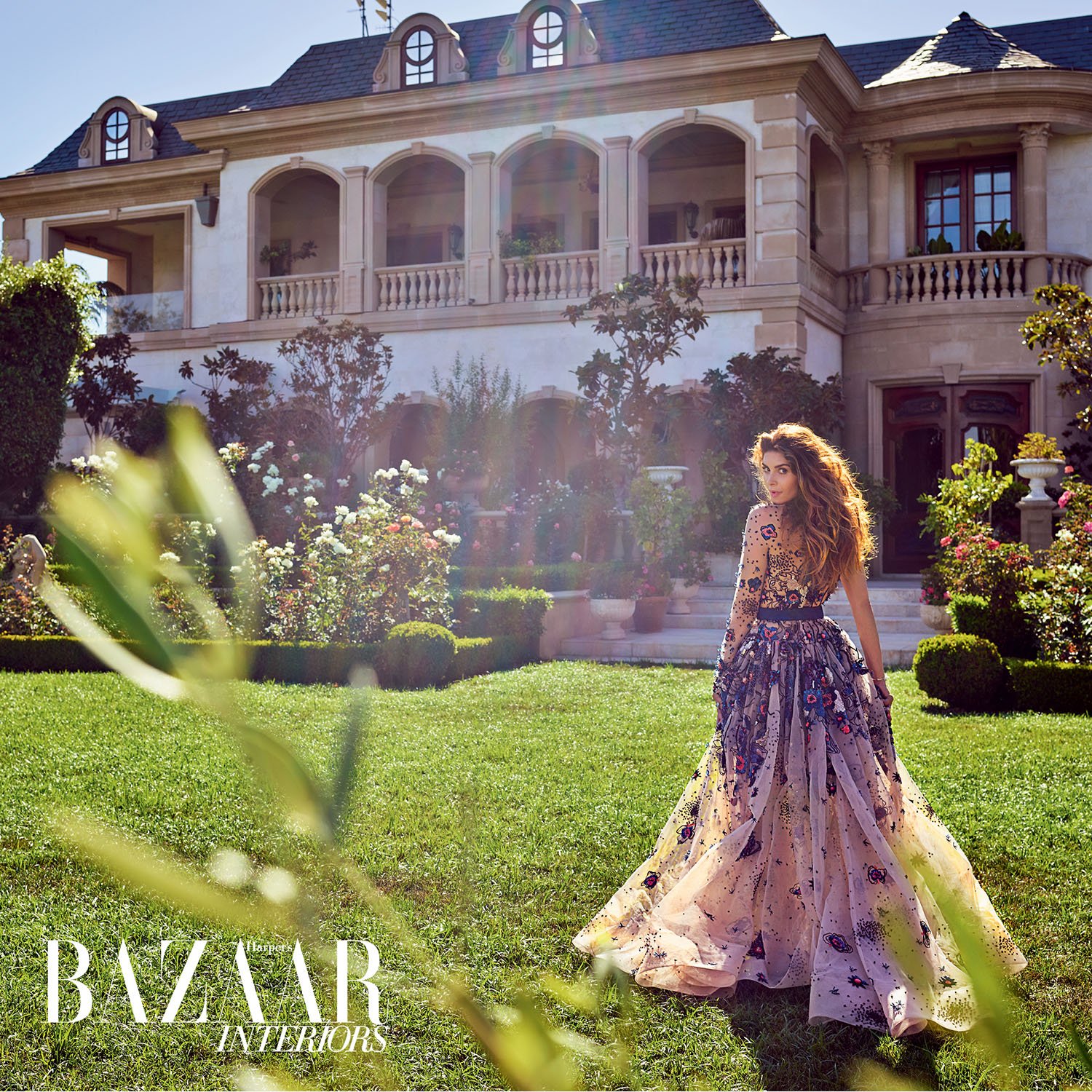 Woman in elegant gown stands in garden of luxury estate. Fashion editorial showcases opulent residential architecture, lush foliage.
