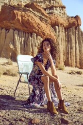 Woman with curly hair in a floral dress seated on a light blue lawn chair in a desert landscape against a rugged sandstone cliff