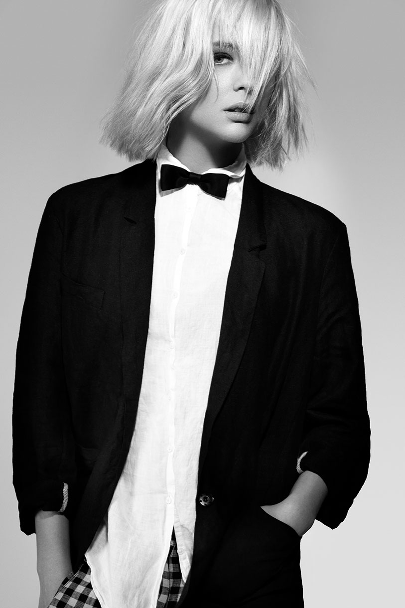 Monochrome portrait of a blonde woman in a black blazer and bow tie against a grey background with a serious expression.