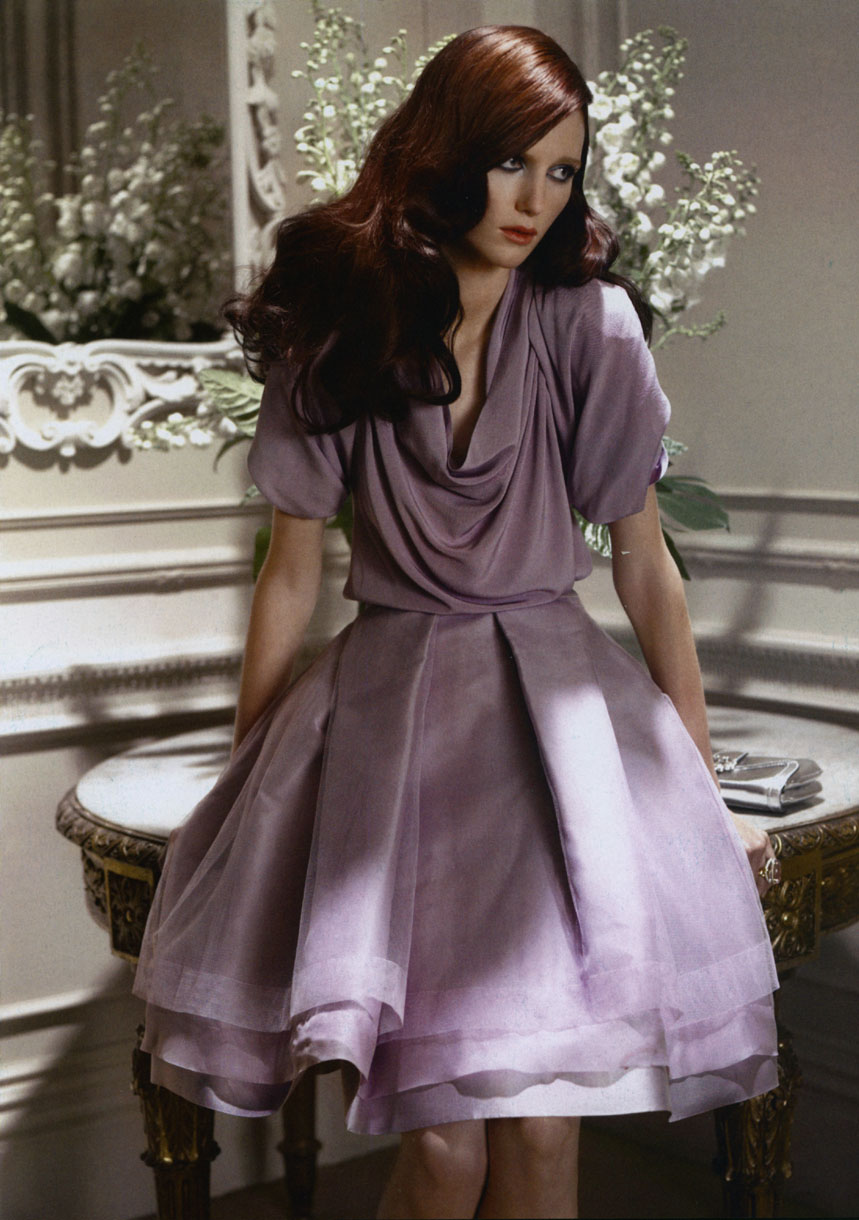 A single brunette female model poses in a lilac dress indoors, in a fashion studio with soft lighting and decorative flowers.