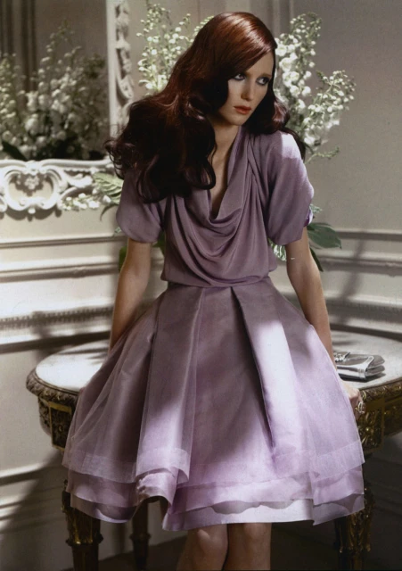 A single brunette female model poses in a lilac dress indoors, in a fashion studio with soft lighting and decorative flowers.