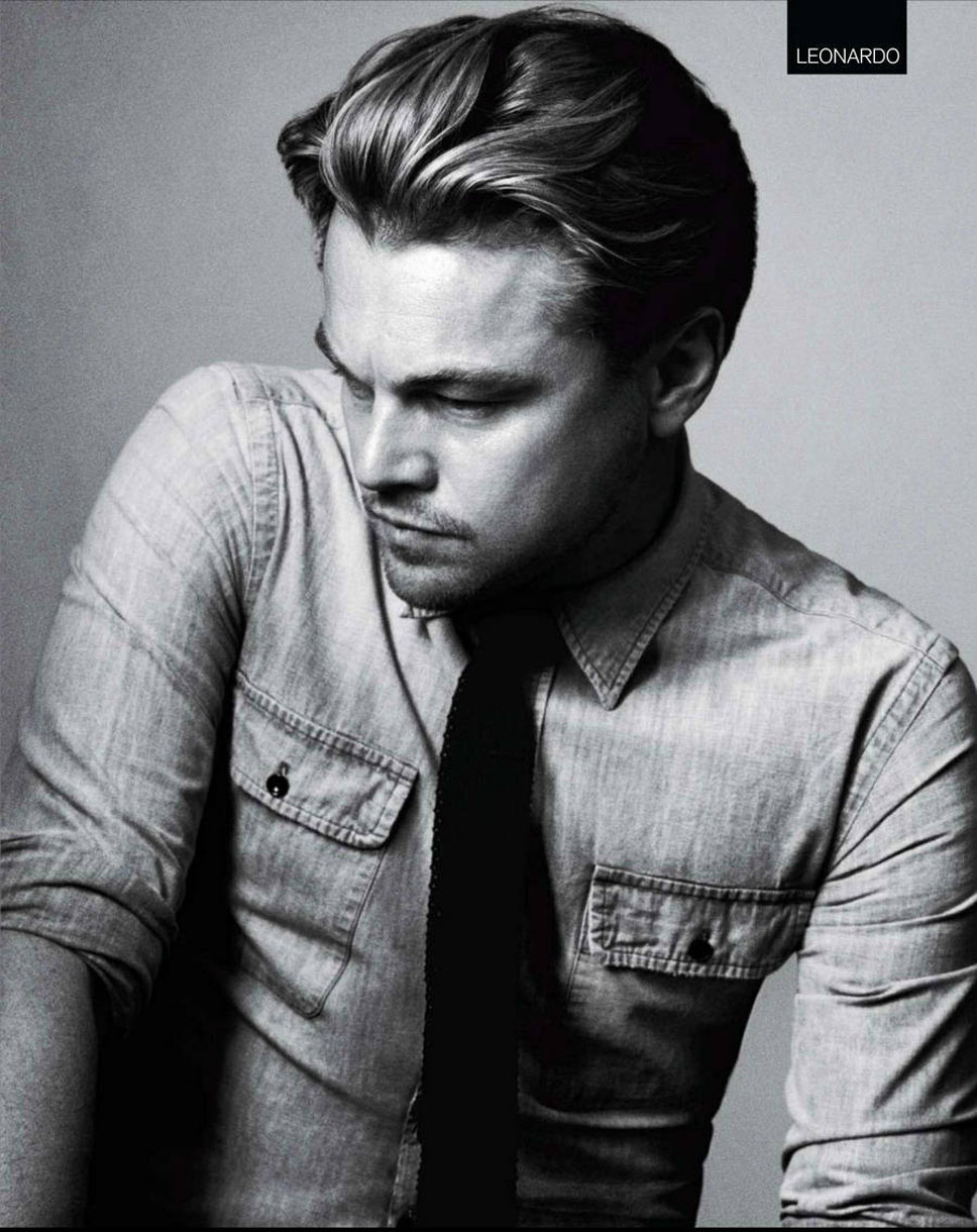 Black and white portrait of Leonardo DiCaprio in a denim shirt and dark tie, gazing downward in a grey gradient background.