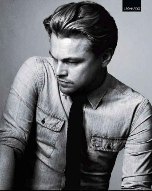 Black and white portrait of Leonardo DiCaprio in a denim shirt and dark tie, gazing downward in a grey gradient background.