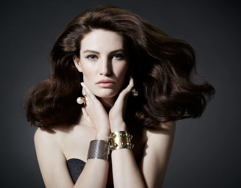 Portrait of a brunette woman with hands near her face wearing jewelry in a studio with a dark background, perfect for beauty advertisement.