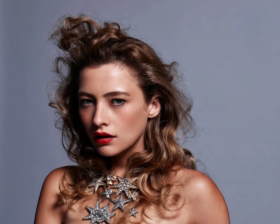 Portrait of a woman with voluminous brown curly hair wearing a star-shaped rhinestone necklace against a grey background.