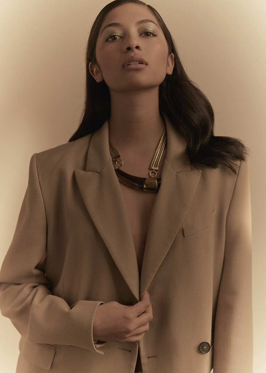 Portrait of an adult woman wearing a beige blazer and a statement necklace with a neutral-toned background in a fashion style image.