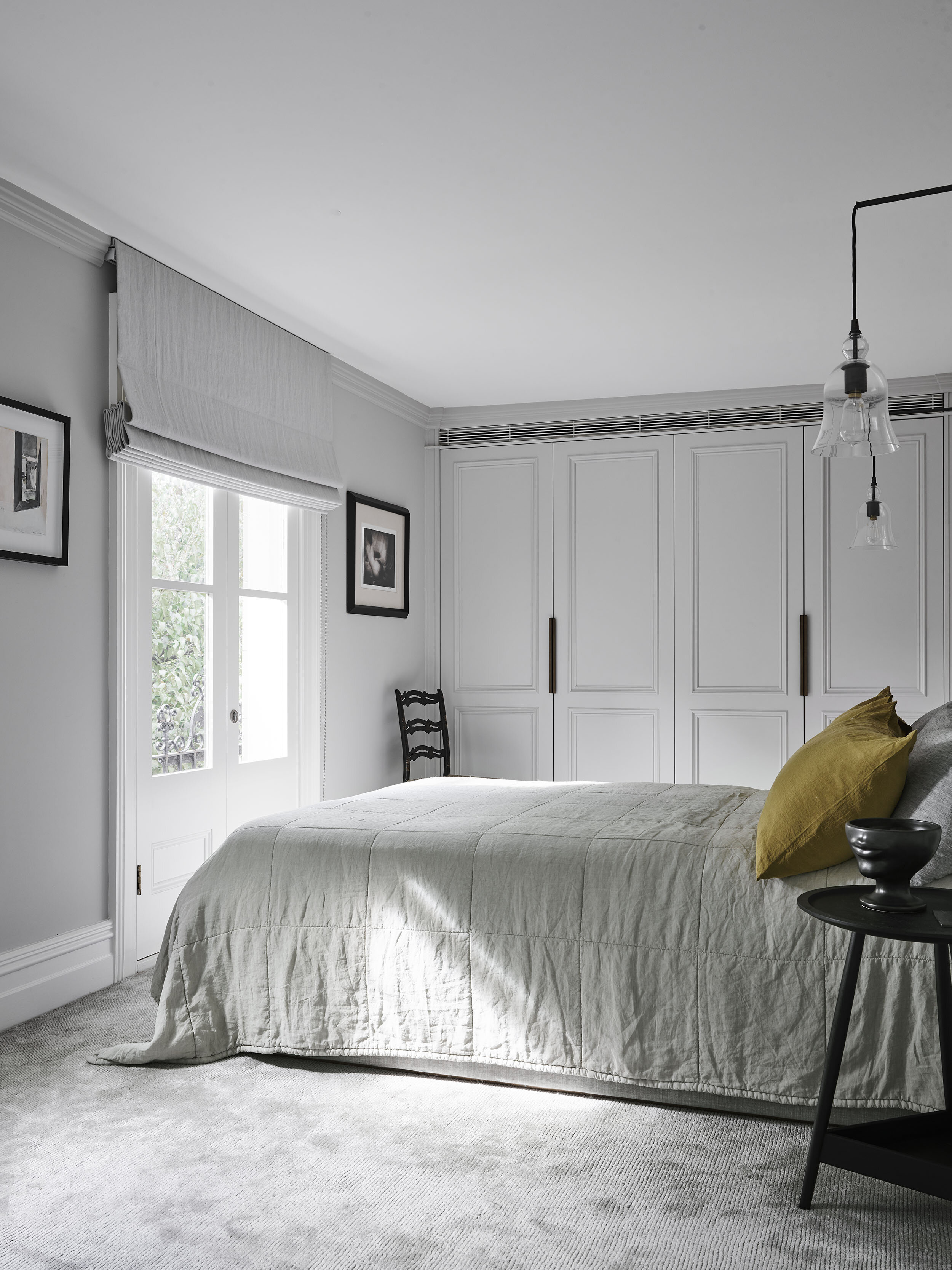Bedroom interior featuring a gray bed with a yellow pillow, a small black side table, and white built-in wardrobes, illuminated by natural light.
