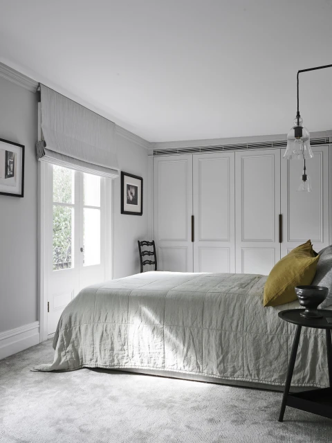 Bedroom interior featuring a gray bed with a yellow pillow, a small black side table, and white built-in wardrobes, illuminated by natural light.