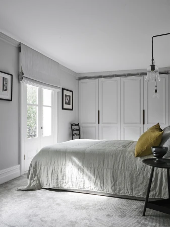 Bedroom interior featuring a gray bed with a yellow pillow, a small black side table, and white built-in wardrobes, illuminated by natural light.