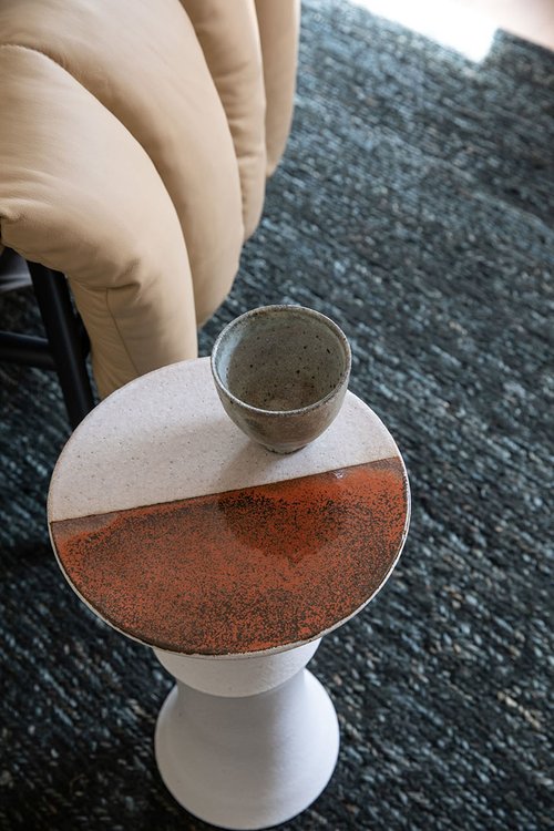 Ceramic teacup sits atop a small round accent table with an orange and white top, complemented by a tufted sofa and textured rug.