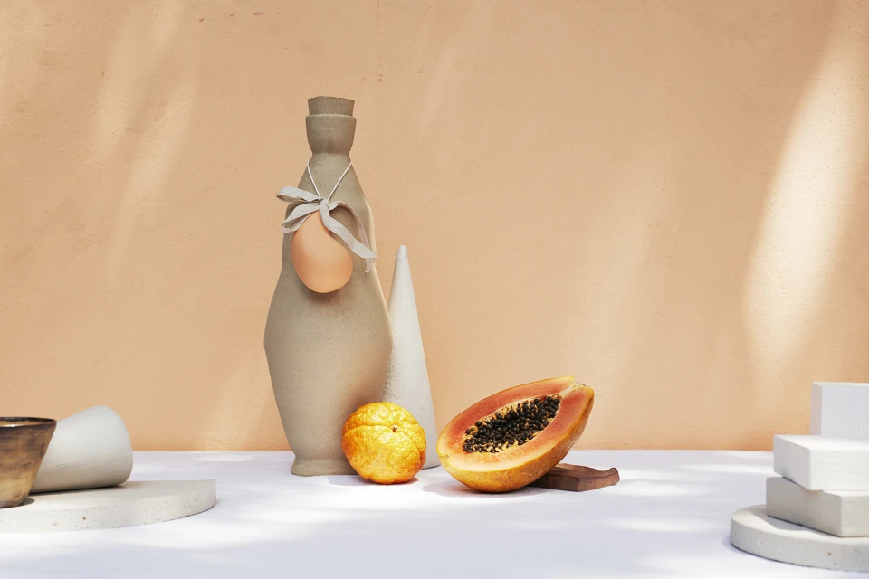 Still life with a decorative bottle, papaya, and orange on a white table against a neutral peach-colored background in a studio setting.