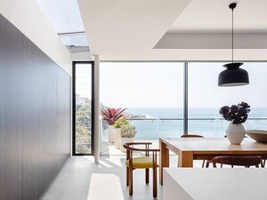 Bright, modern dining area with black accent wall, skylight, wooden table, potted plants on a balcony overlooking a scenic ocean view.