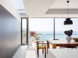 Bright, modern dining area with black accent wall, skylight, wooden table, potted plants on a balcony overlooking a scenic ocean view.