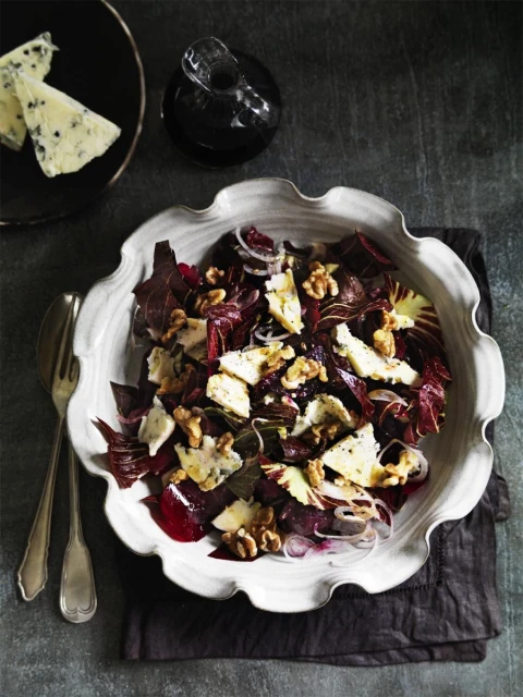 Overhead shot of radicchio salad in a white bowl with Roquefort cheese, walnuts, and vinaigrette on a gray surface with silverware.
