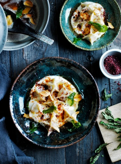 Overhead shot of blue plates containing manti dumplings in yogurt sauce garnished with mint and sumac on a wooden table.