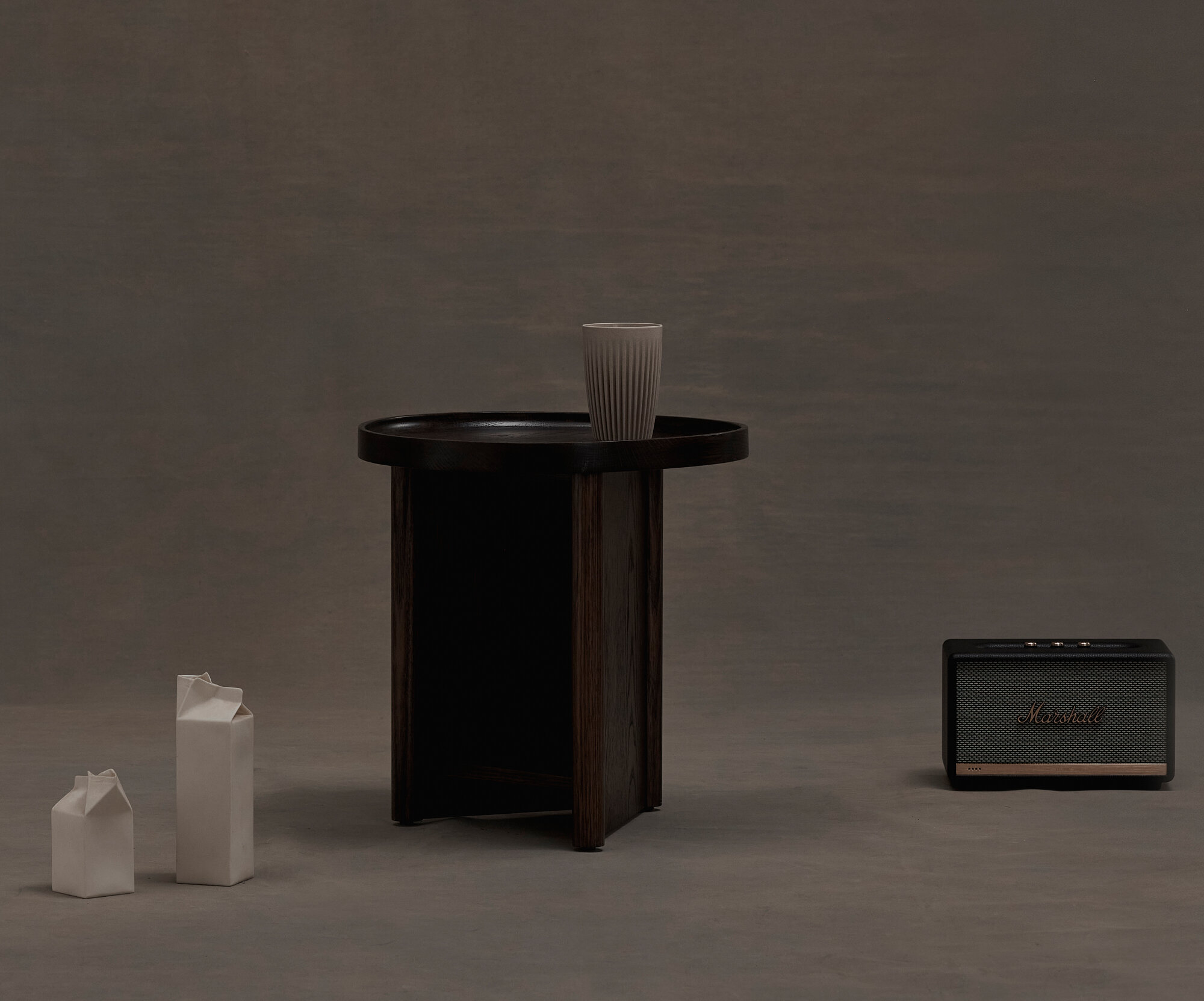 Still life shot of a side table with a cup, two milk carton sculptures, and a speaker against a grey backdrop in studio lighting.