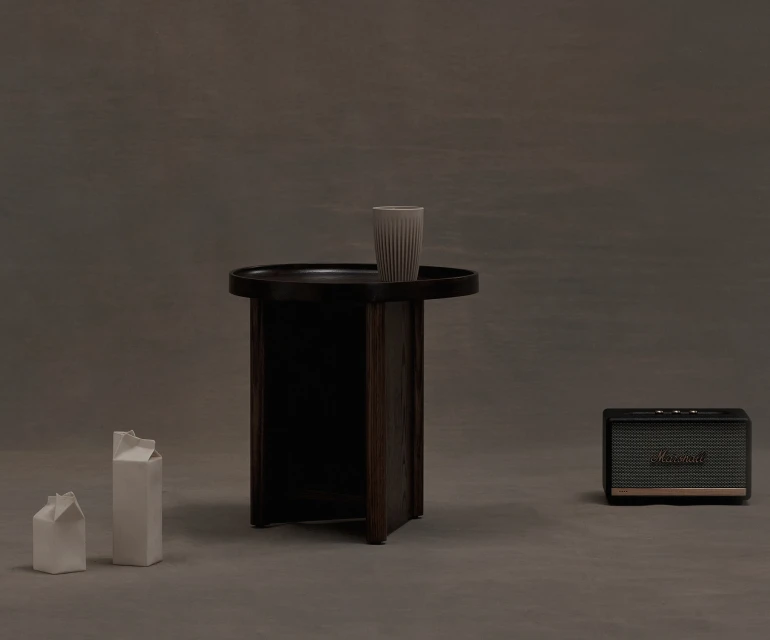 Still life shot of a side table with a cup, two milk carton sculptures, and a speaker against a grey backdrop in studio lighting.