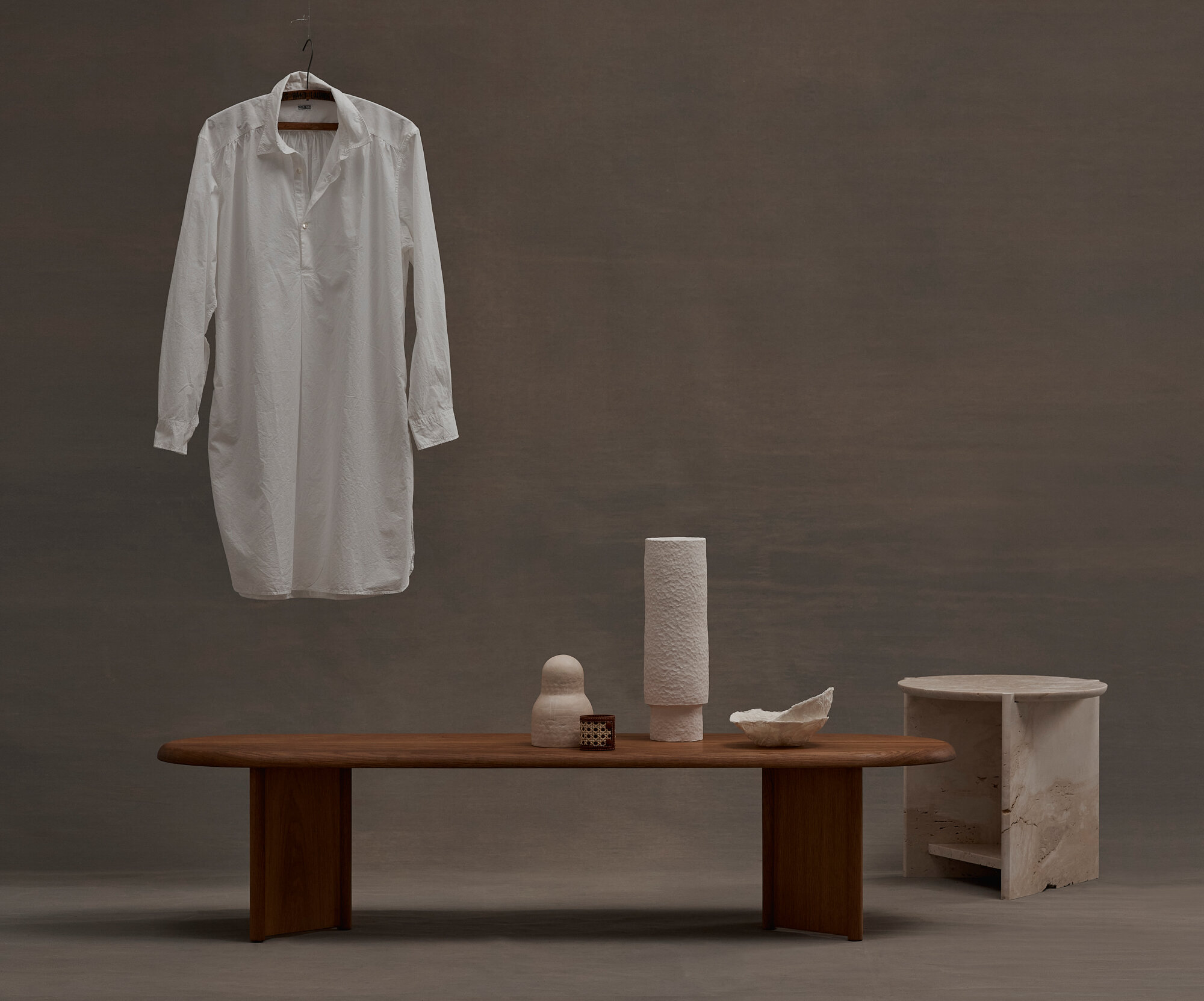 Minimalist fashion photography featuring a white shirt dress on a hanger, displayed with ceramic art pieces on a wooden table against a grey backdrop.