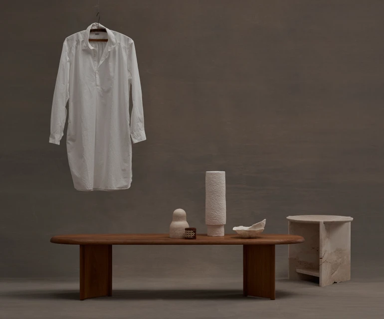 Minimalist fashion photography featuring a white shirt dress on a hanger, displayed with ceramic art pieces on a wooden table against a grey backdrop.