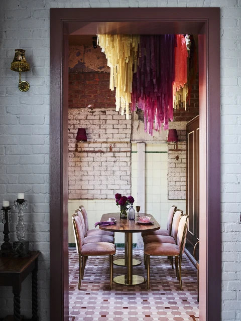 Dining room with decorative streamers hanging from the ceiling, set with a dark wood table, pink velvet chairs and brick walls.