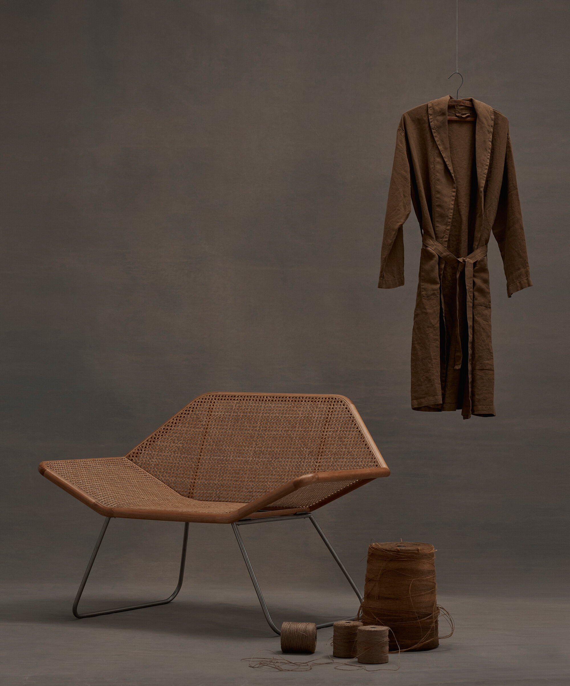 A minimalist scene showcases a modern rattan chair with metal legs, a brown robe on a hanger, a woven basket, and twine rolls on a grey surface.