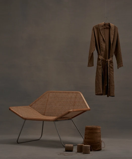 A minimalist scene showcases a modern rattan chair with metal legs, a brown robe on a hanger, a woven basket, and twine rolls on a grey surface.