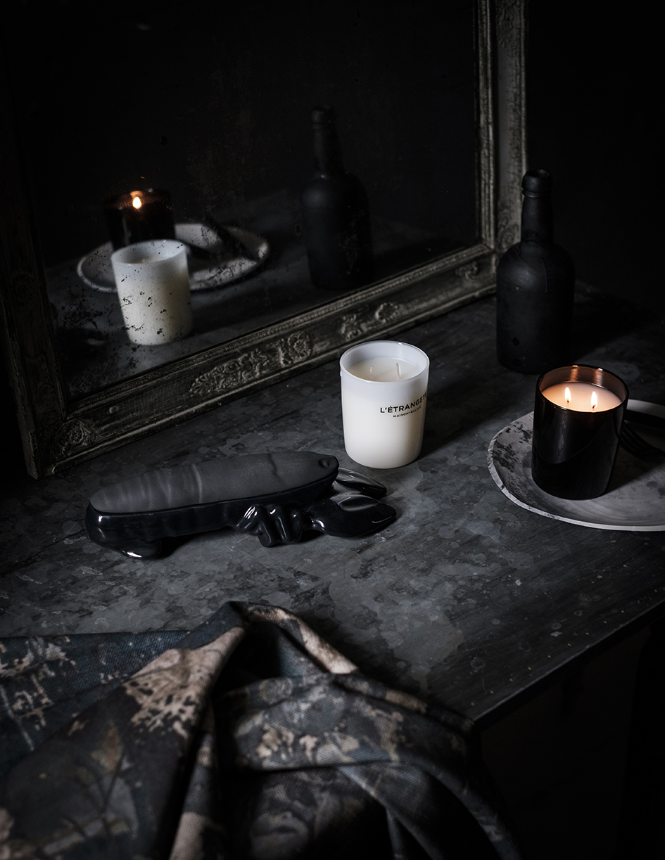 Dark, moody still life composition featuring lit candles, a sculptural object, and draped fabric on a textured gray surface.