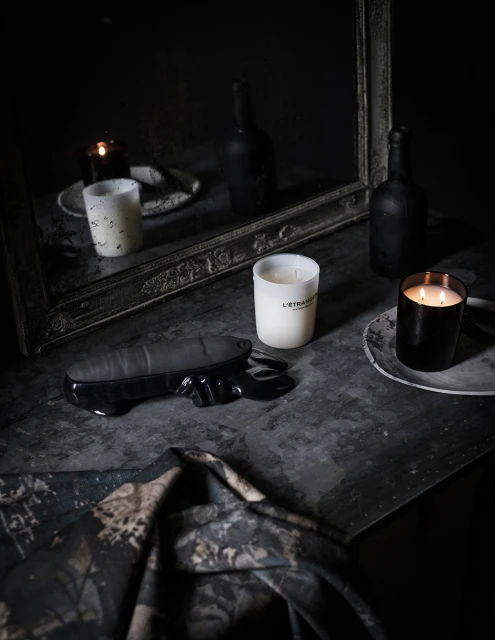 Dark, moody still life composition featuring lit candles, a sculptural object, and draped fabric on a textured gray surface.