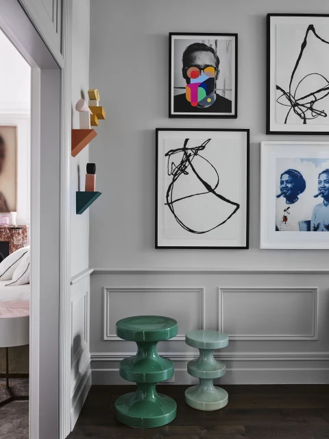 Interior of a hallway with gray walls and hardwood flooring, featuring a pair of green sculptural stools, framed wall art, and geometric wall decor.