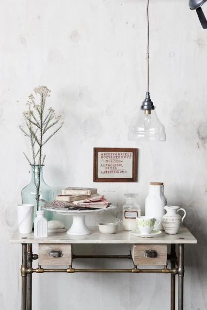 Still life of a desk with books, bottles, flowers, and a pendant light in a rustic, vintage-inspired setting against a textured wall.