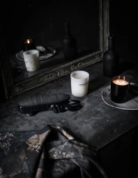 Dark moody still life with scented candles, glass bottles, and a textured cloth on a rustic surface.