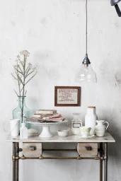 Interior styling by Claire Delmar featuring a rustic table with ceramics, glassware, and a framed sampler on a whitewashed wall.