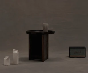 Dark wood side table with a ribbed ceramic cup, two white paper cartons, and a vintage speaker on a grey seamless backdrop