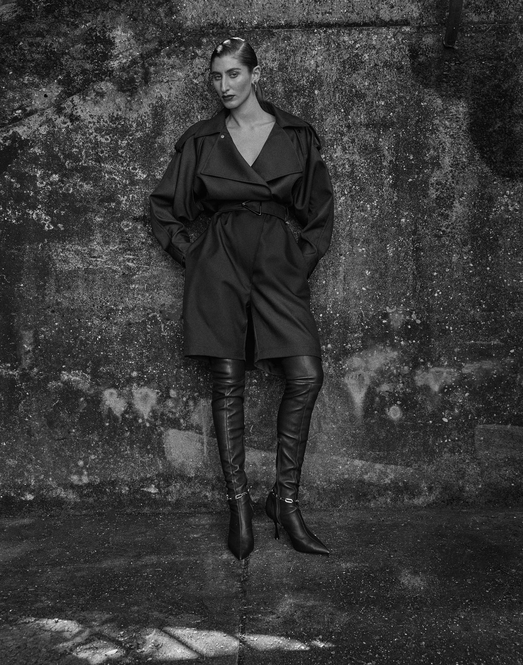 Black and white studio shot of adult female fashion model in a dark belted trench coat and thigh-high boots posing against a textured wall.