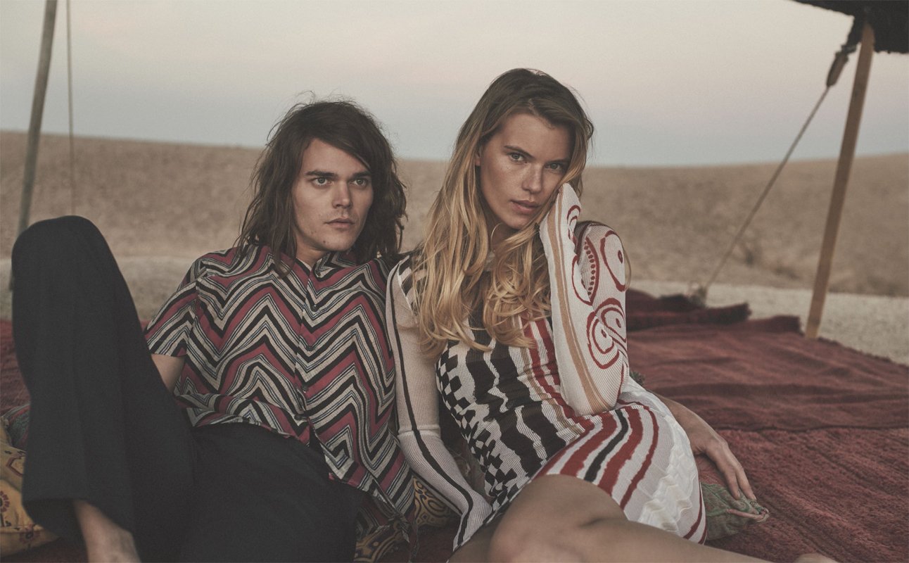 Fashion shot featuring an adult male and female posing on a carpet in a desert-like setting with earth tones and geometrical patterns.