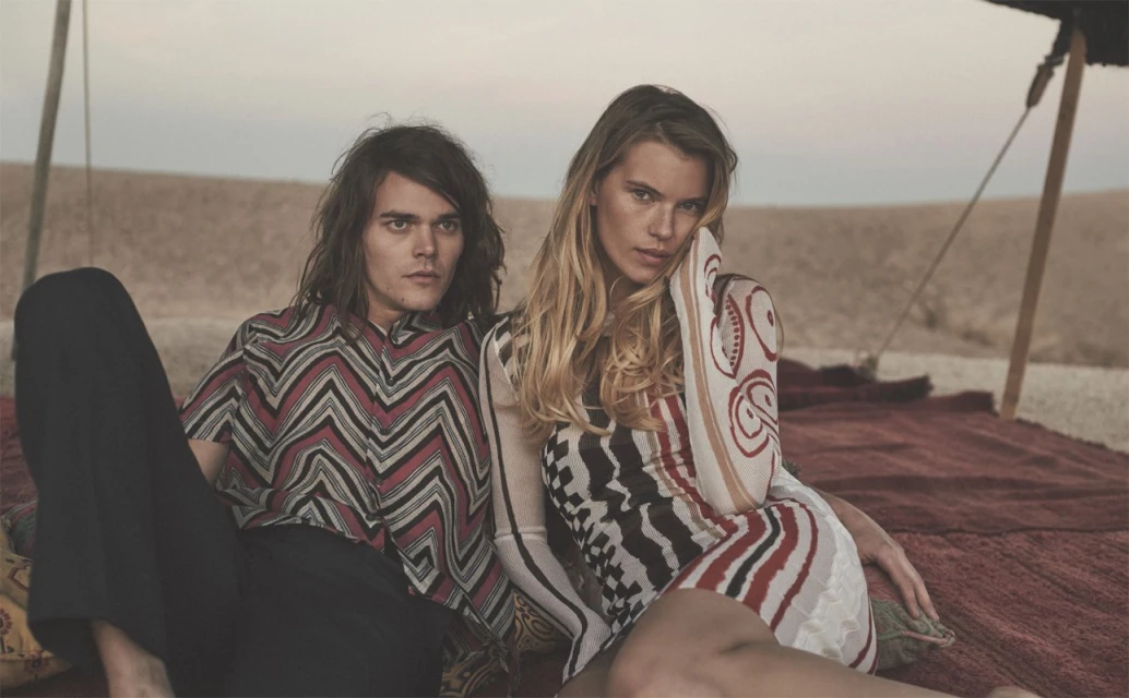 Fashion shot featuring an adult male and female posing on a carpet in a desert-like setting with earth tones and geometrical patterns.