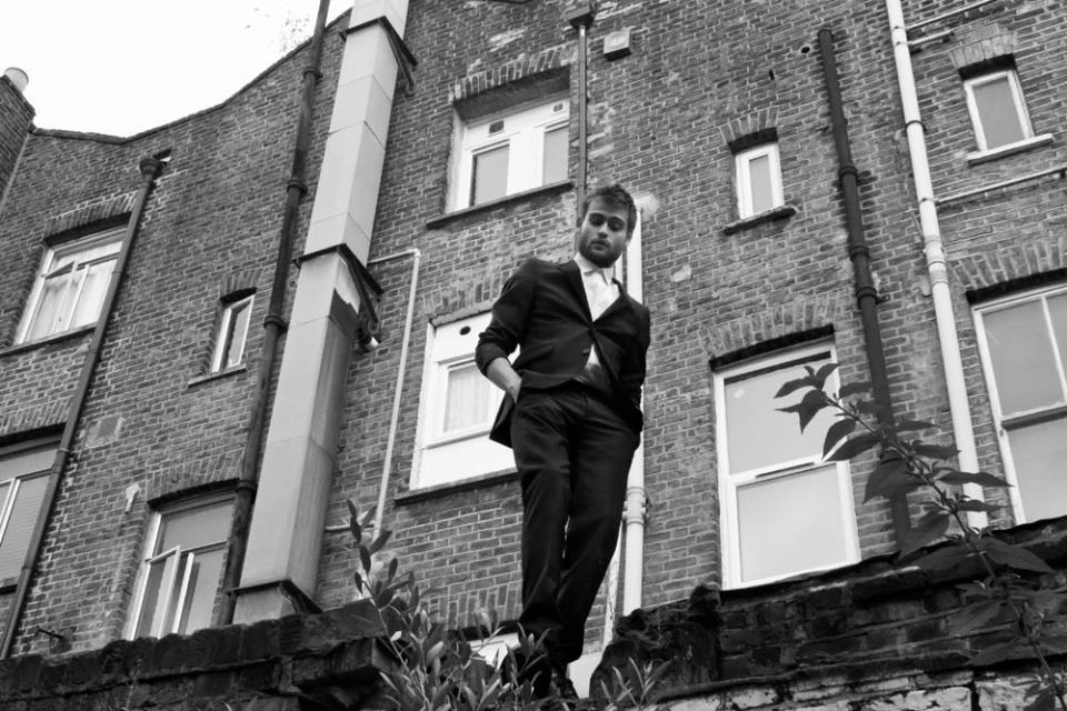 Low angle portrait of an adult male in a suit standing on a brick ledge against a brick building, captured in black and white.