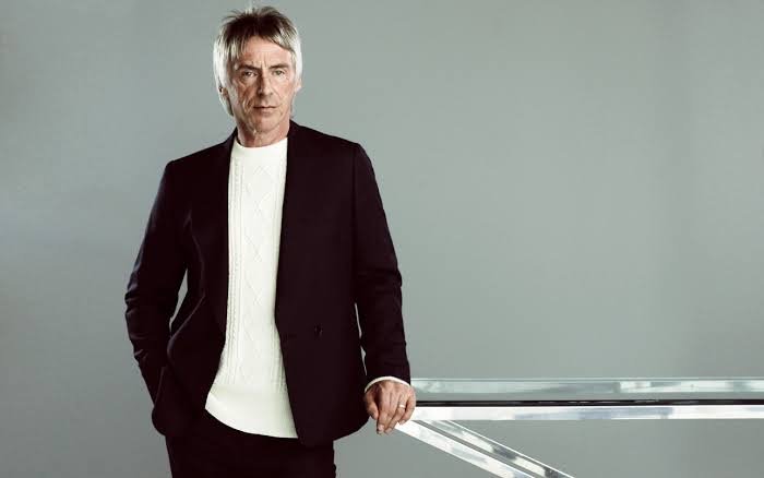 A mature adult man in a black suit jacket and white patterned sweater stands indoors against a gray background, posing confidently.
