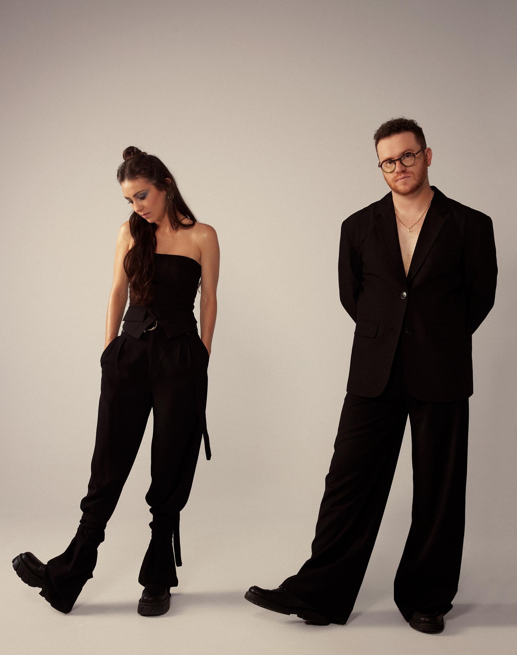 Studio shot of a woman in a black tube top and pants next to a man in an unbuttoned black suit. Both are in fashionable attire.