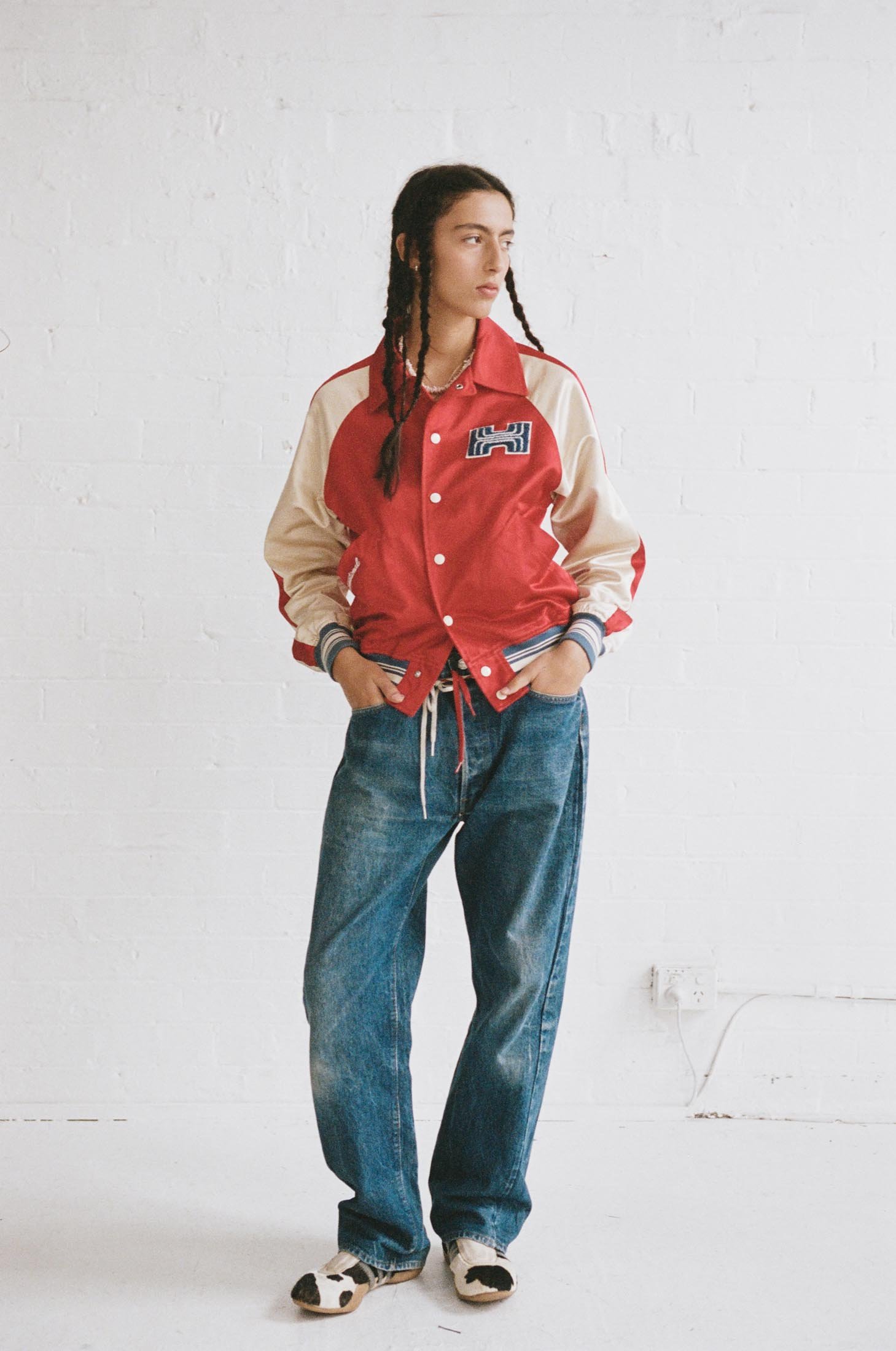 Full-length shot of a young adult woman in a red and beige varsity jacket and jeans, posing in front of a white wall in a studio.