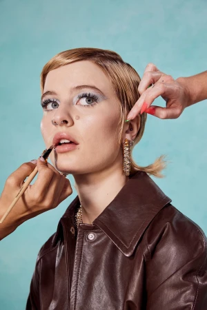 Close up beauty portrait of a woman with short blonde hair wearing a brown leather jacket while hands apply makeup and style her hair.