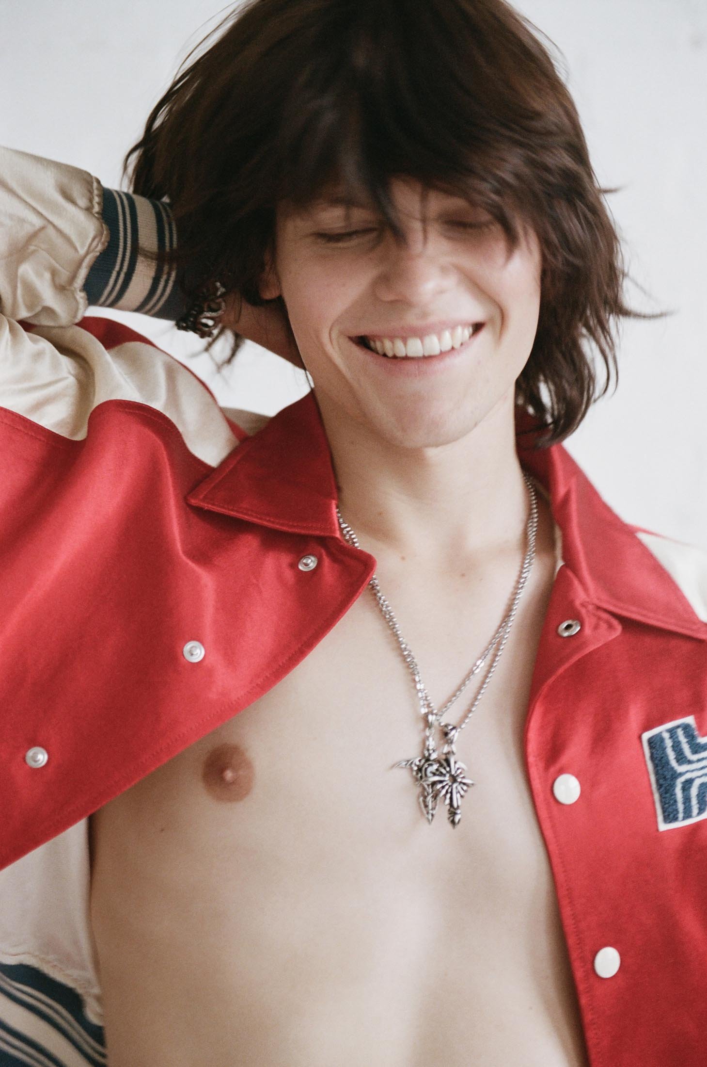 Close-up of a smiling, fair-skinned young adult with shaggy brunette hair, wearing an open red varsity jacket and necklace, against a white wall.