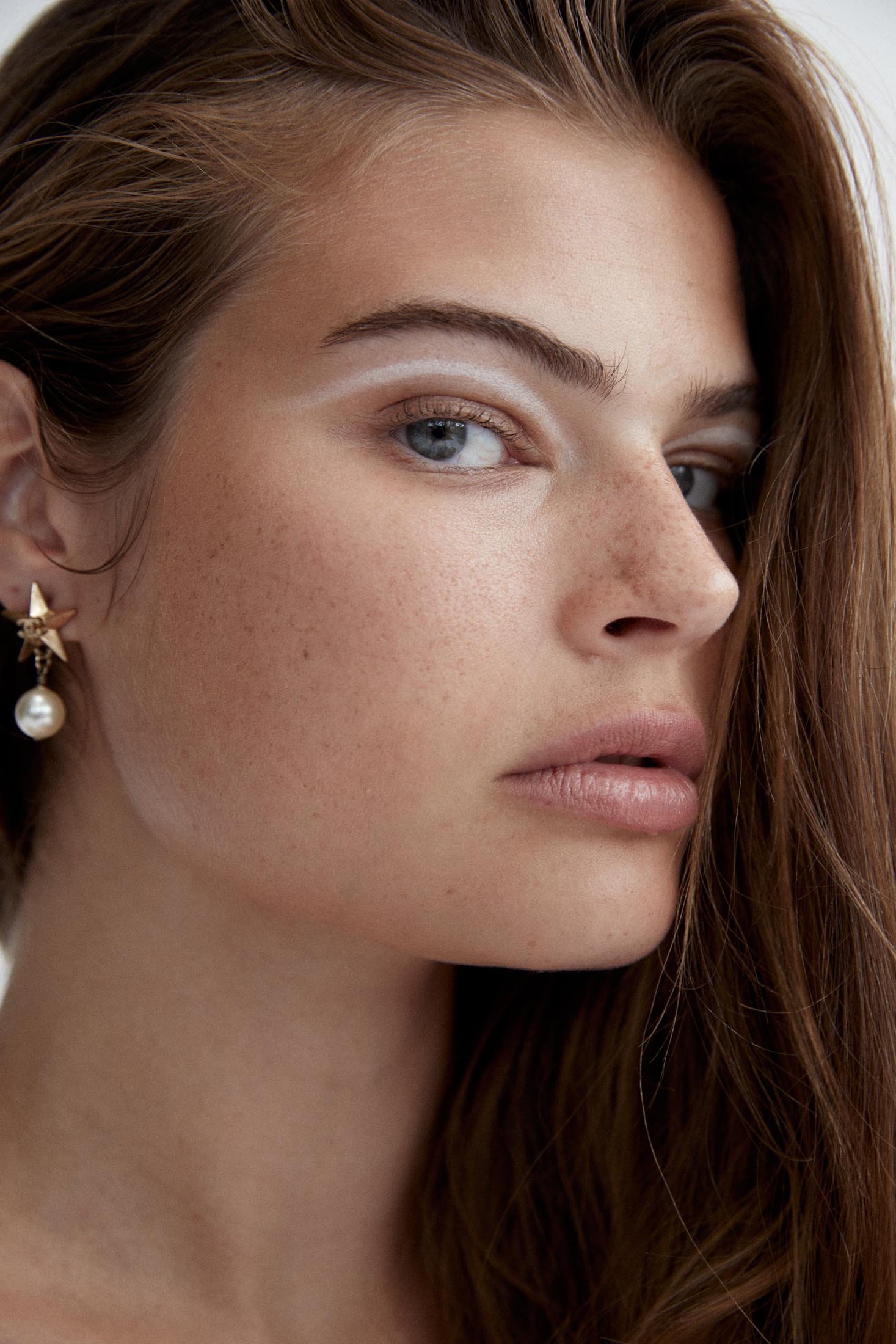Close-up portrait of a young adult woman with long brown hair, pearl earrings, and creative white makeup around her eyes in a neutral setting.
