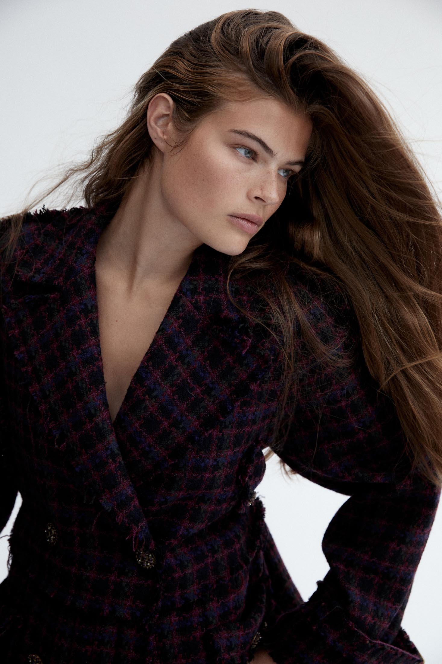 Adult female wearing a plaid tweed double-breasted blazer with flowing brown hair in a studio fashion shot against a white background.