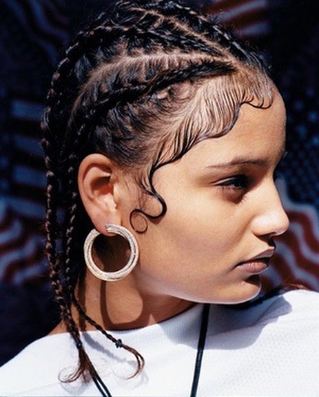 Portrait of a young woman with braided hair and hoop earrings, posing in front of a blurred background with colors of the American flag.