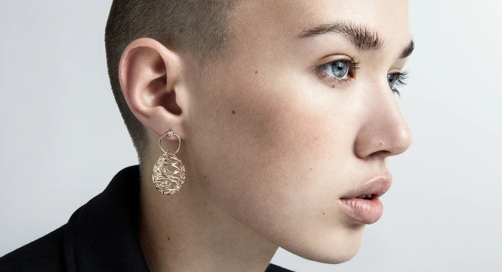 Close-up profile of a young adult woman with a buzzcut wearing a decorative gold earring against a white background in a studio setting.