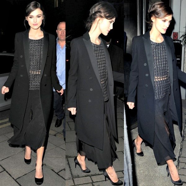 Keira Knightley wearing a black double-breasted coat, a patterned top, and flowing black pants. She is seen on a city street at night.